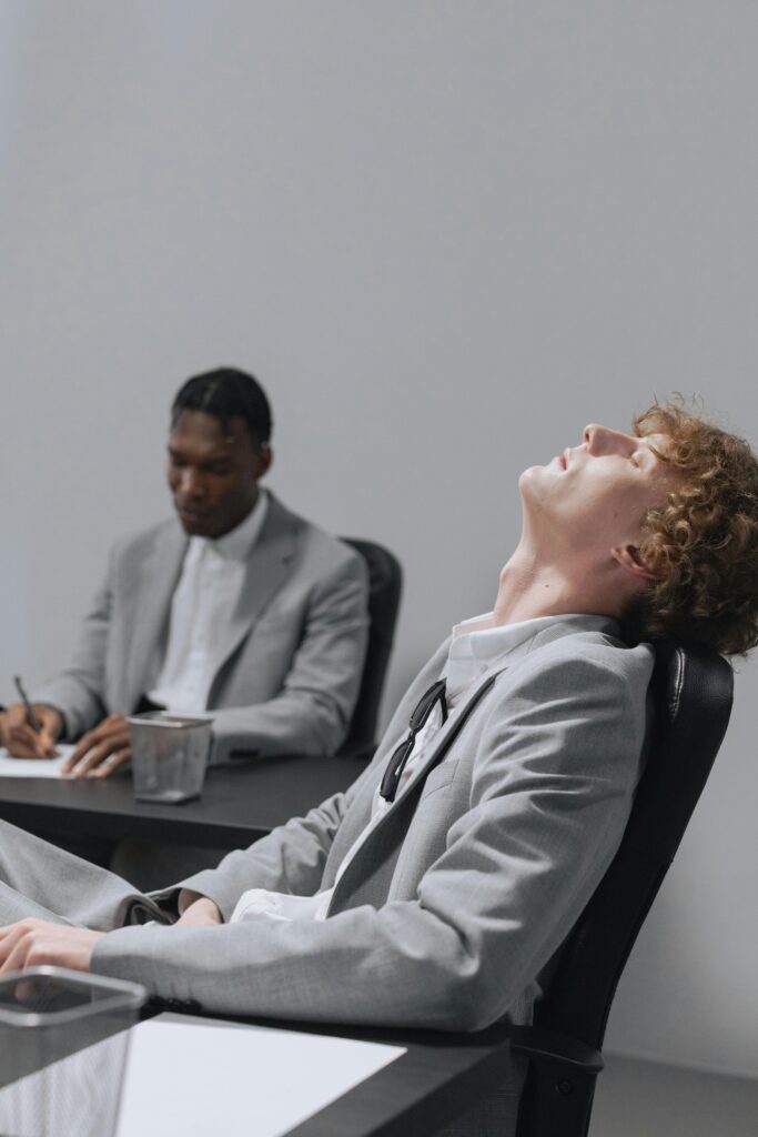 Two young professionals in gray suits sitting at desks, one appearing bored, indoors.